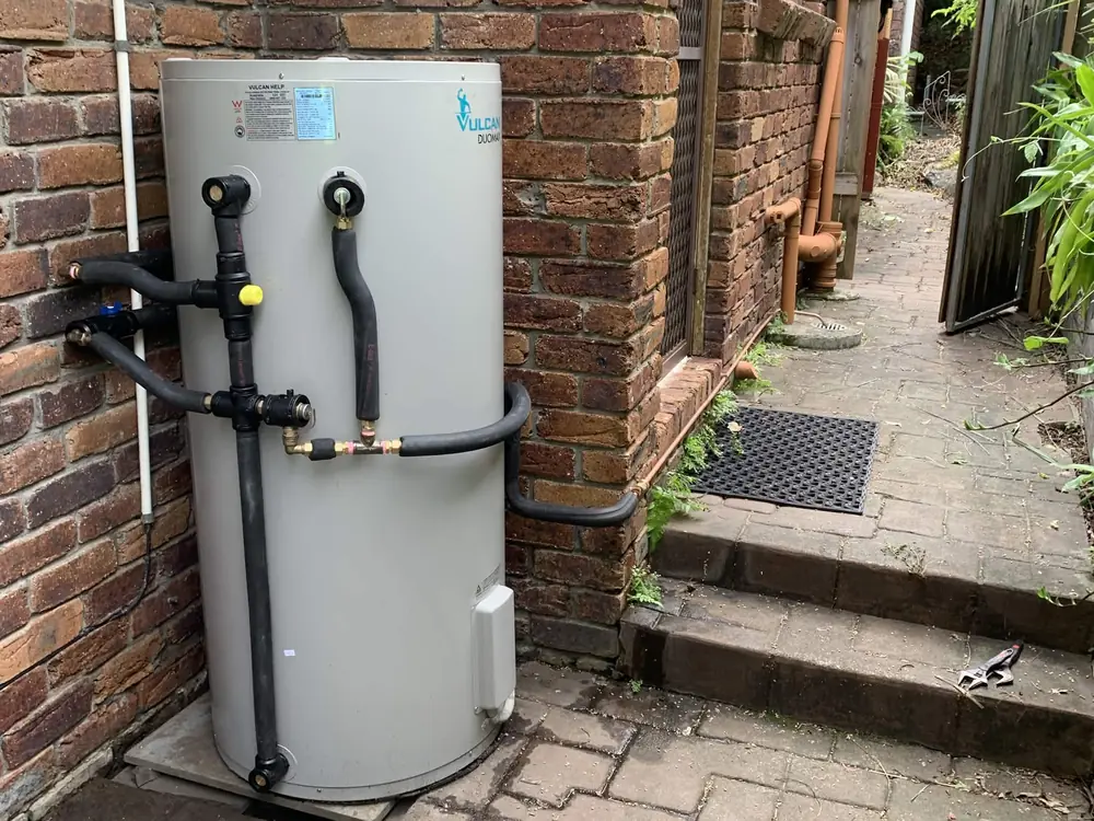 After — a rusted, mossy electric hot water system swapped for a new Vulcan unit with tidy new pipework